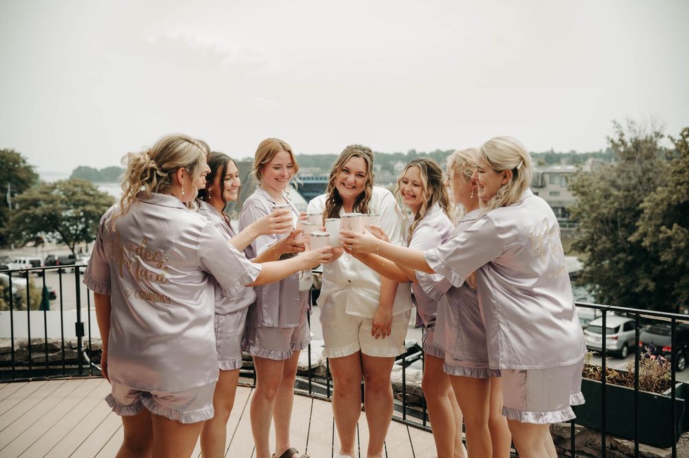 Bride and bridesmaids in matching satin pajamas toast champagne glasses together on a rooftop balcony.