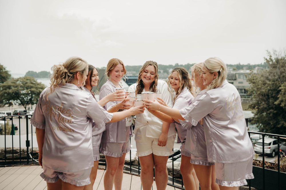 Bride in white robe and bridesmaids in silver satin sets clink champagne glasses on a scenic waterfront balcony.