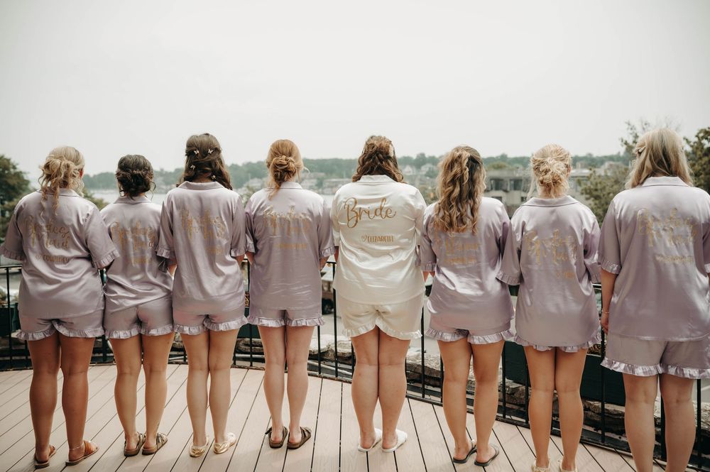 Bride and bridesmaids show off matching satin pajamas from behind, with bride wearing a white Bride-embroidered robe.