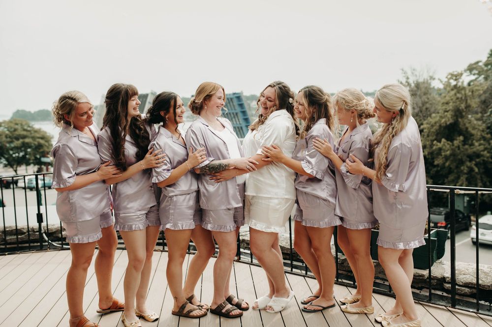 Bridesmaids in matching satin robes hugging the bride on a rooftop deck before the wedding.