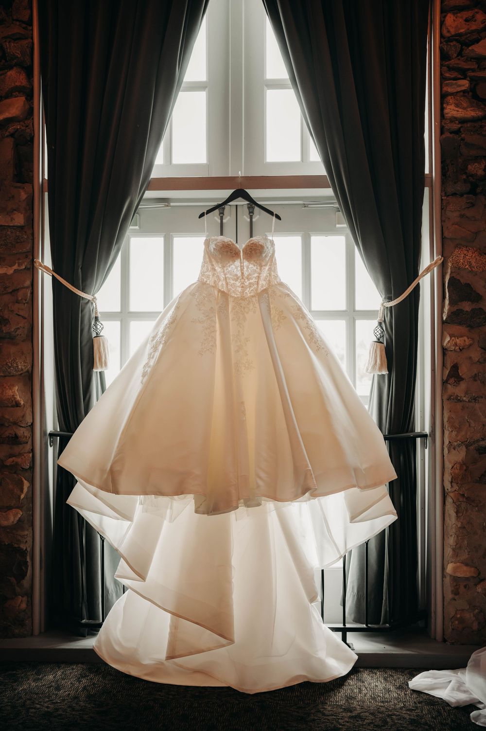 Elegant strapless ball gown wedding dress hanging in a window with dramatic curtains.
