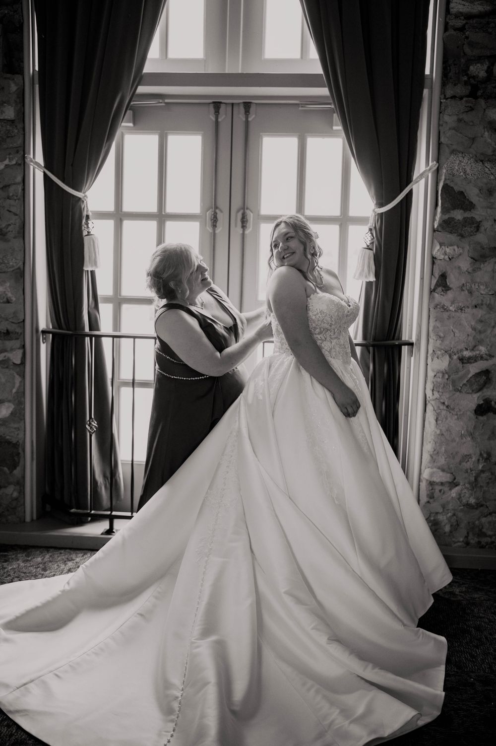 Black and white photo of mother helping bride into her wedding gown by a window.