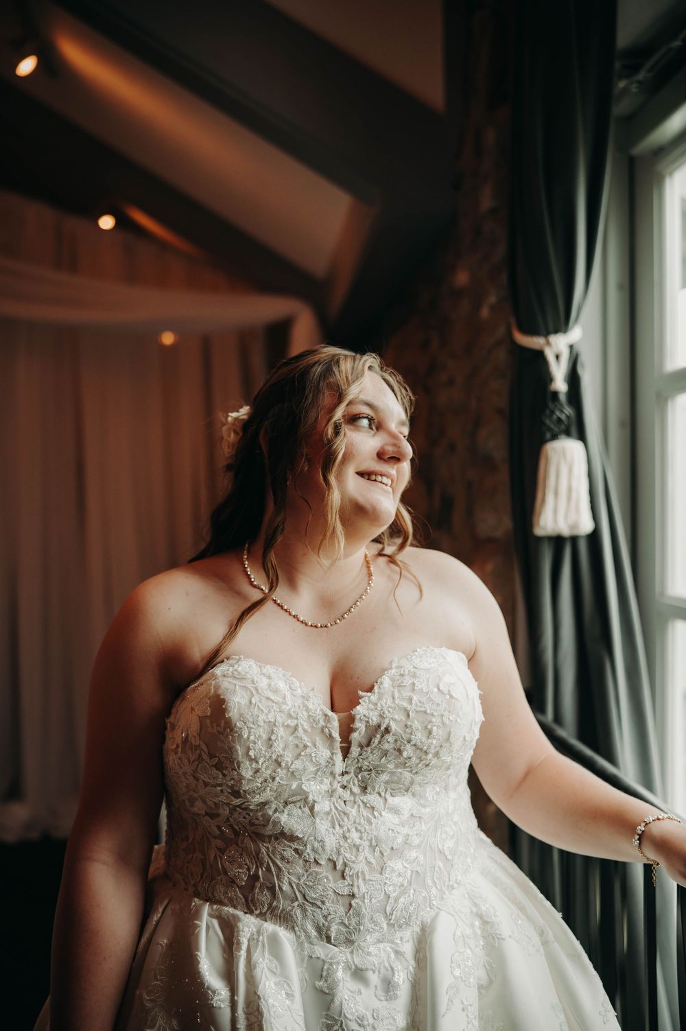Radiant bride in a lace strapless wedding gown smiling softly by a draped window.