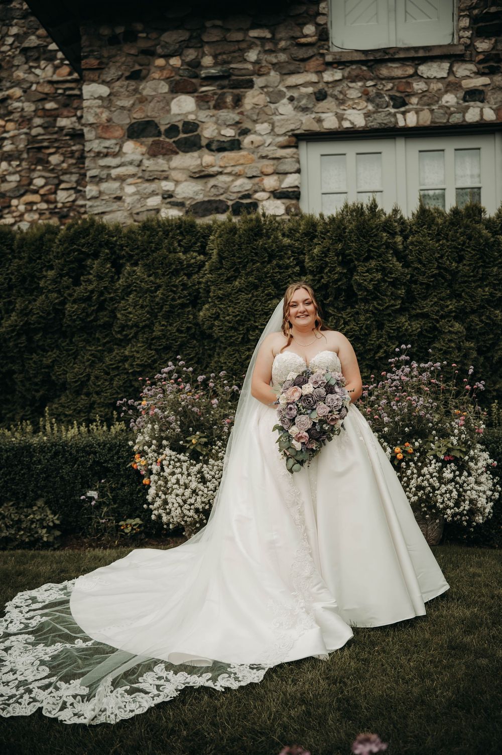 Bride in strapless ball gown with lace veil holding purple bouquet outdoors.