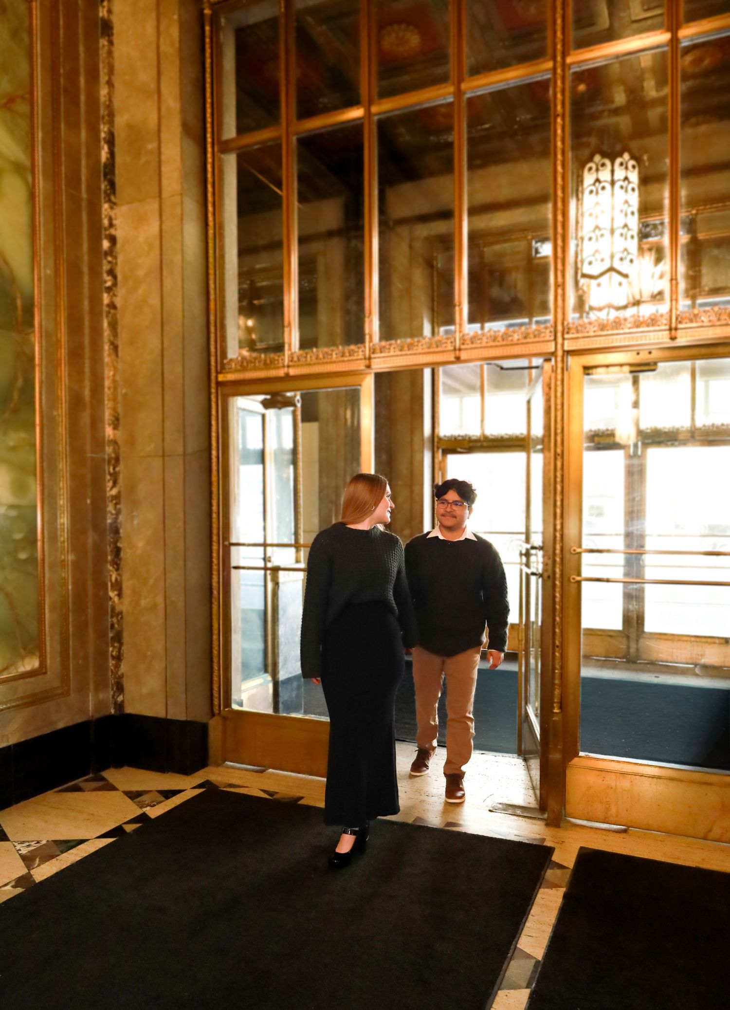 A couple stands in conversation near glass doors in an elegant vintage building interior.
