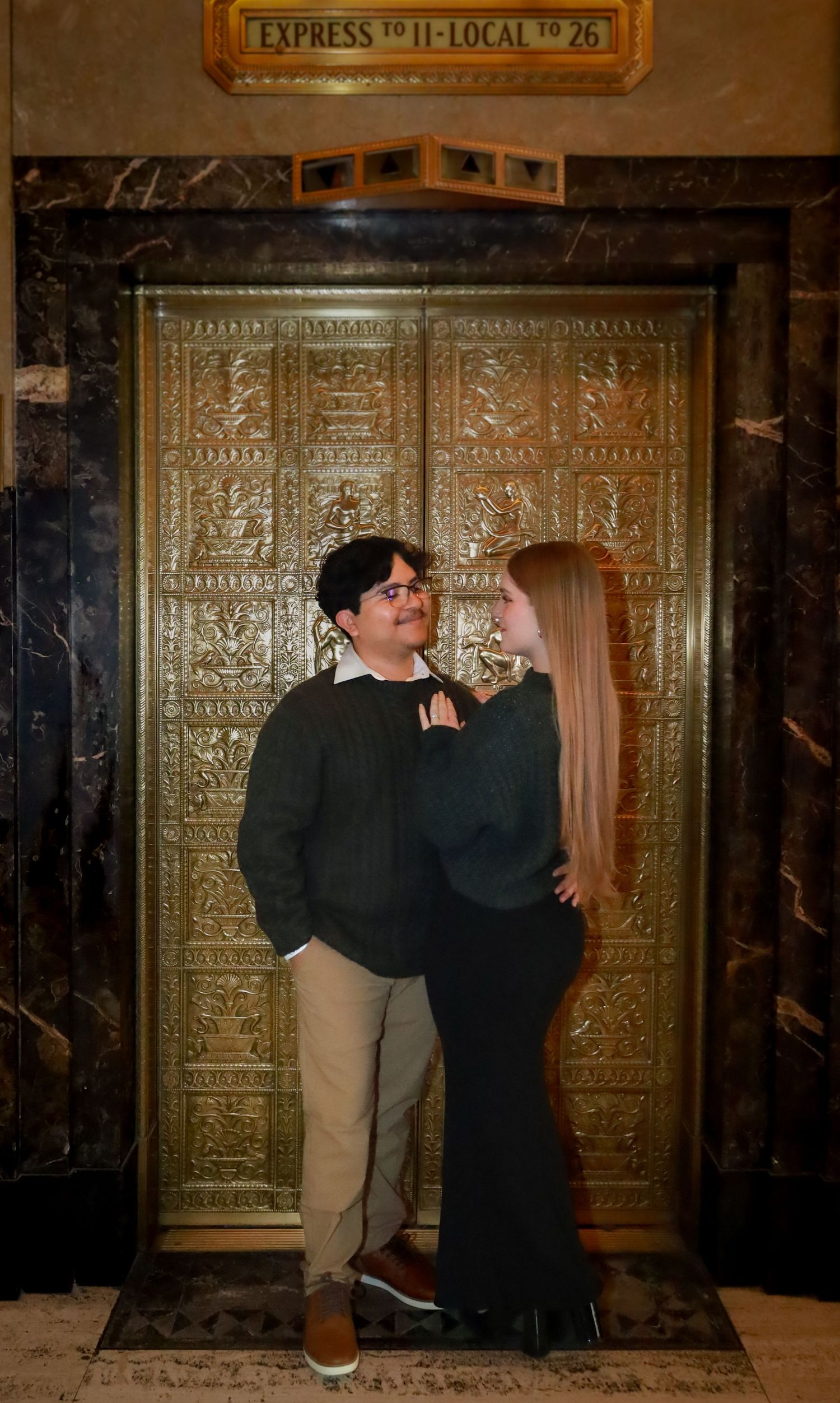 A couple poses in front of an ornate gold elevator door with art deco details.