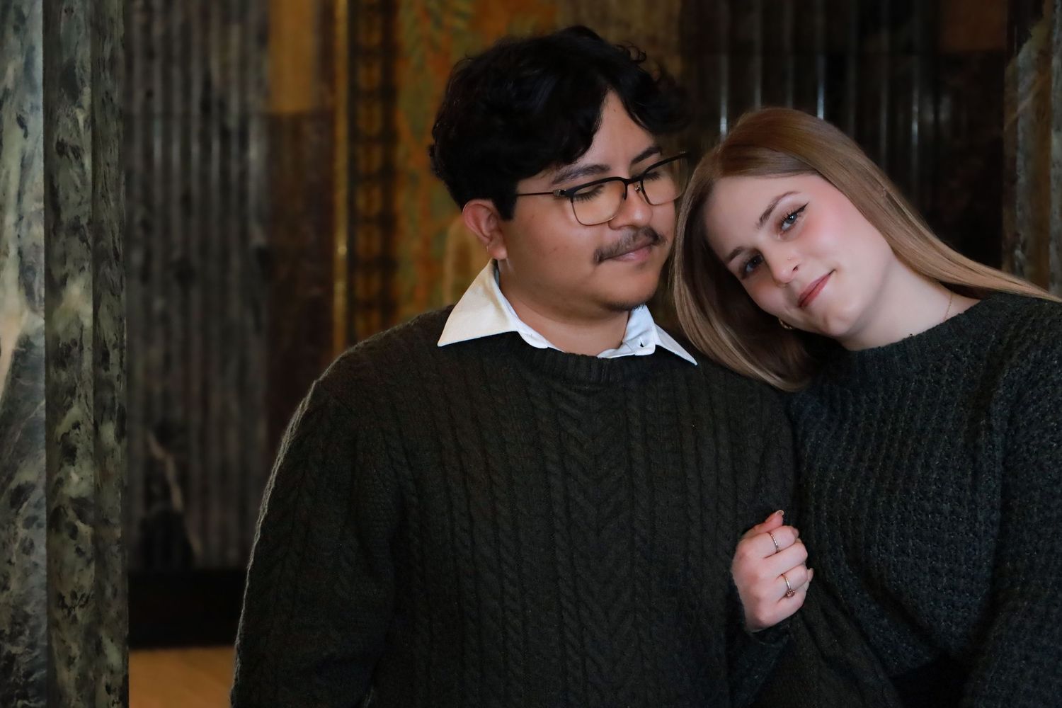 A couple in matching dark sweaters embraces in front of a vintage wall.