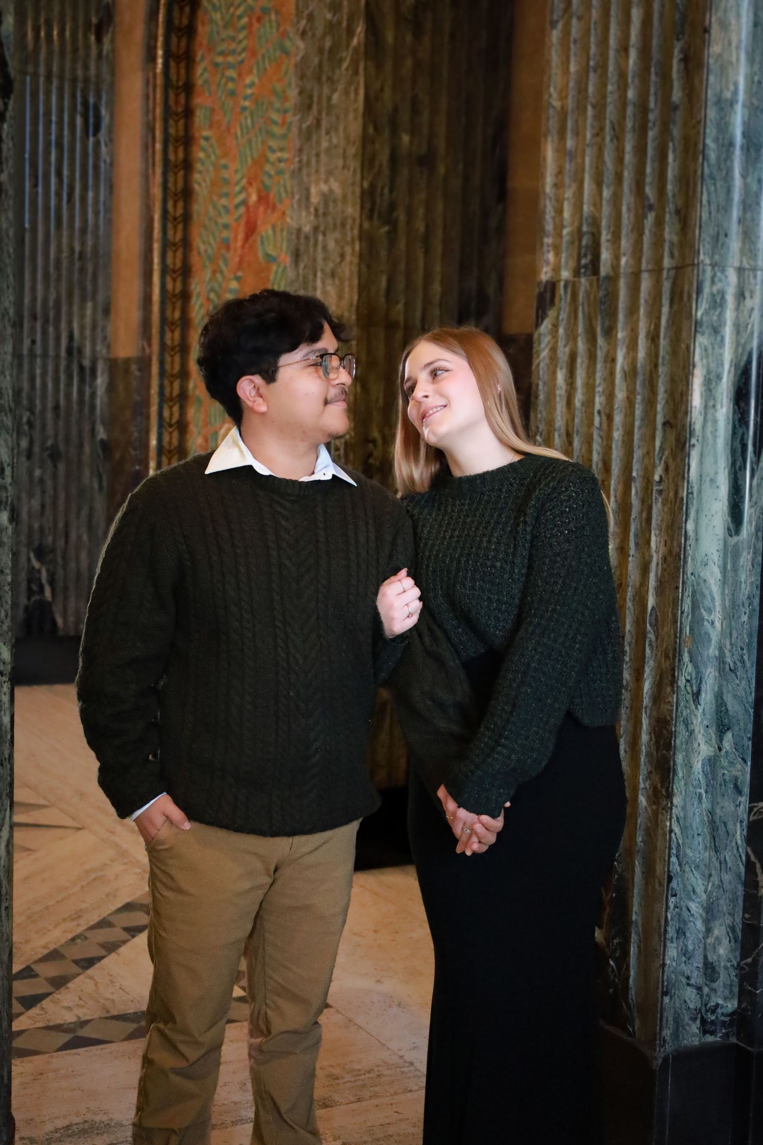 A couple in casual winter wear shares a loving glance in an ornate hallway.
