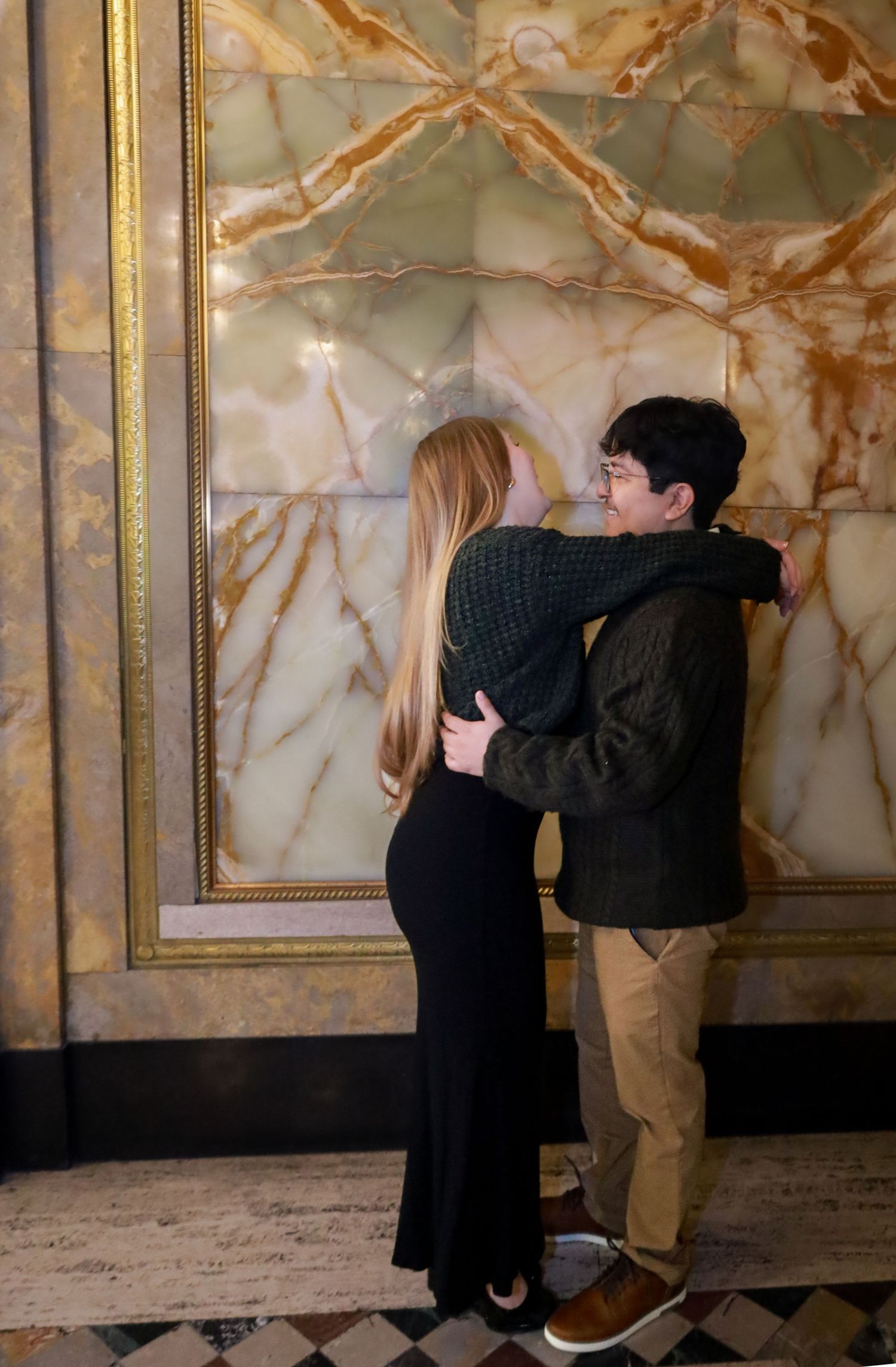 A couple embraces in front of a luxurious marble wall with gold accents.