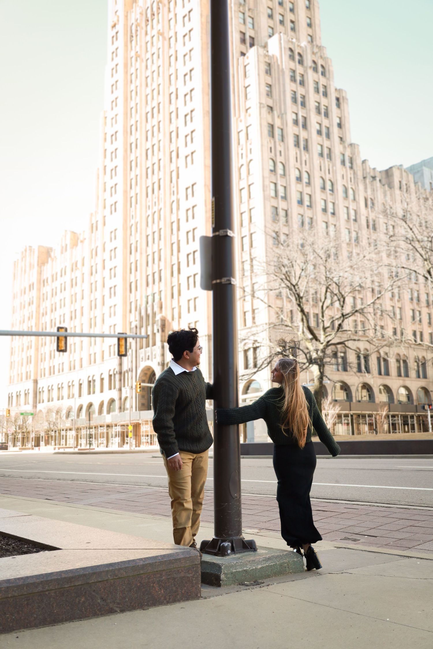 A couple poses playfully near a light pole with an art deco building in the background.