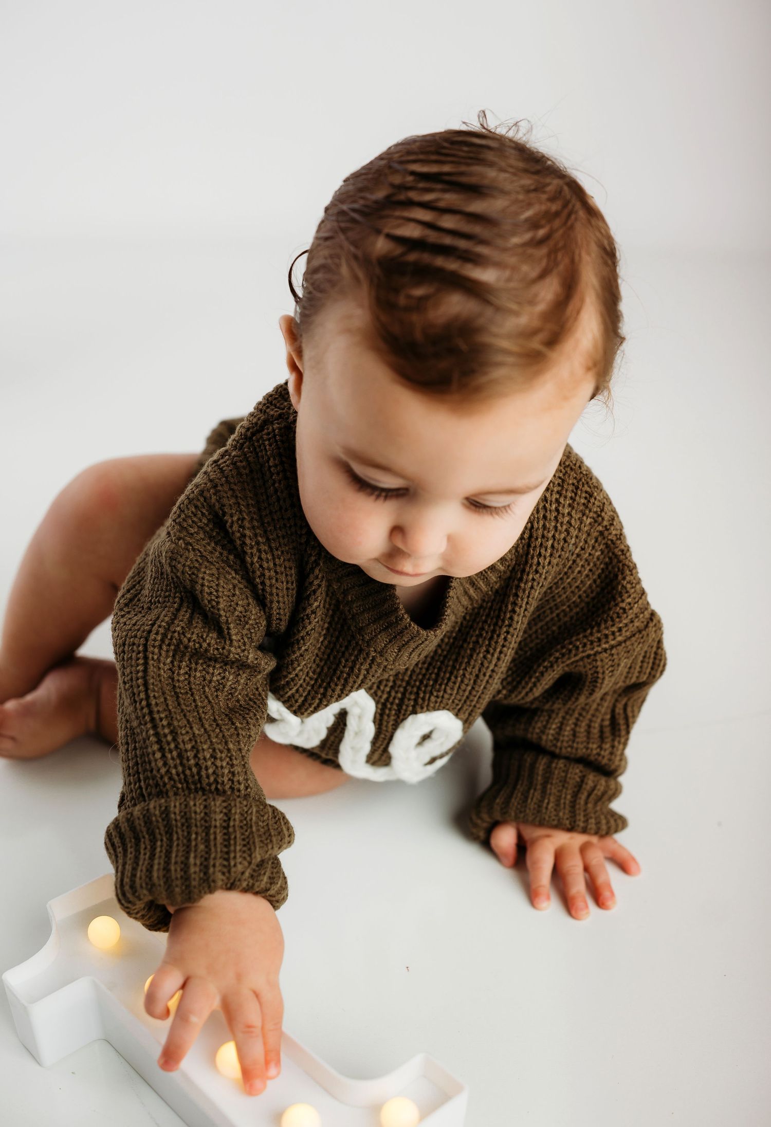 Baby in brown knit sweater crawling on white surface reaching toward glowing lights.