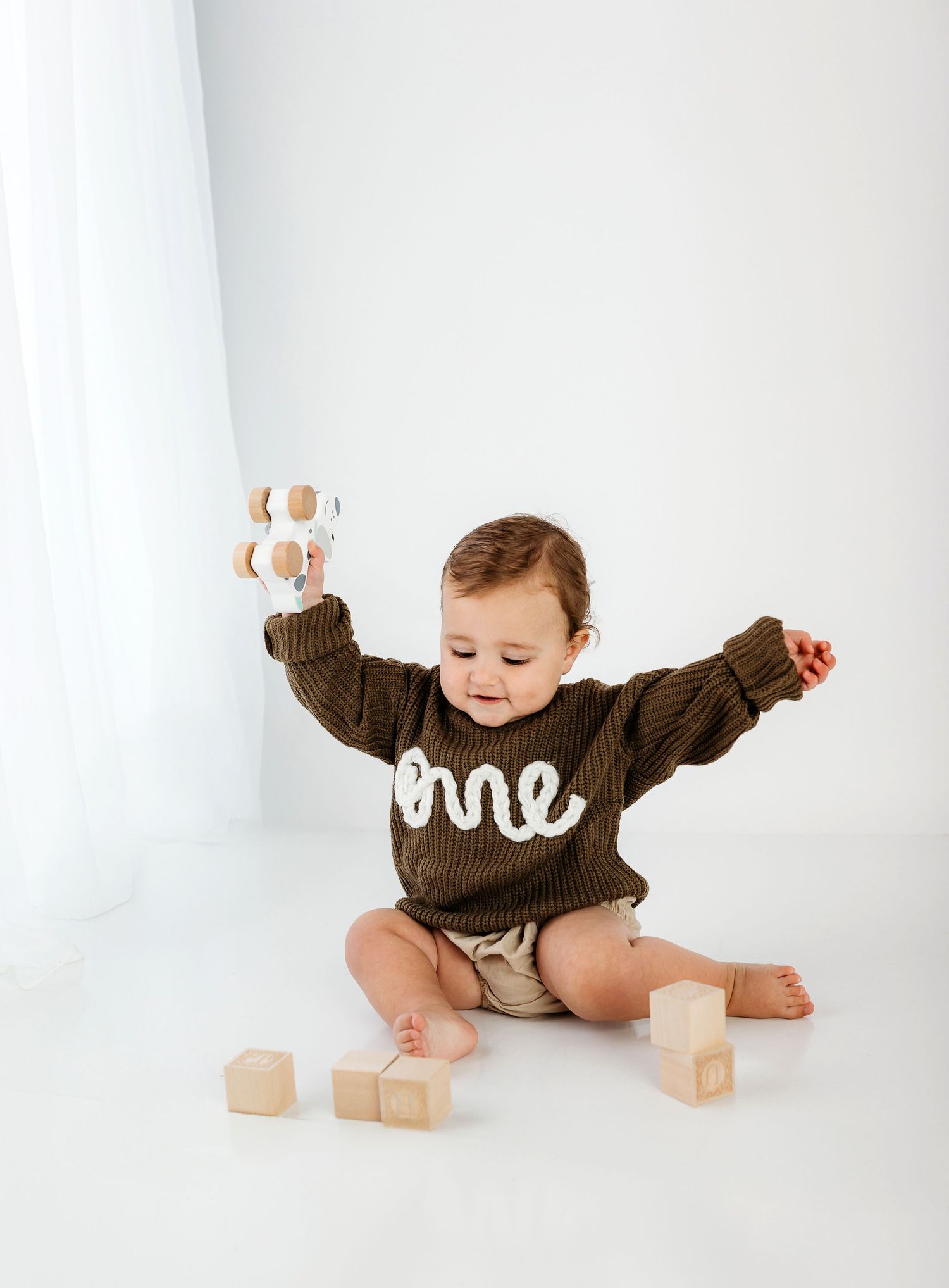 A baby in a brown sweater plays with wooden alphabet blocks on a white background.