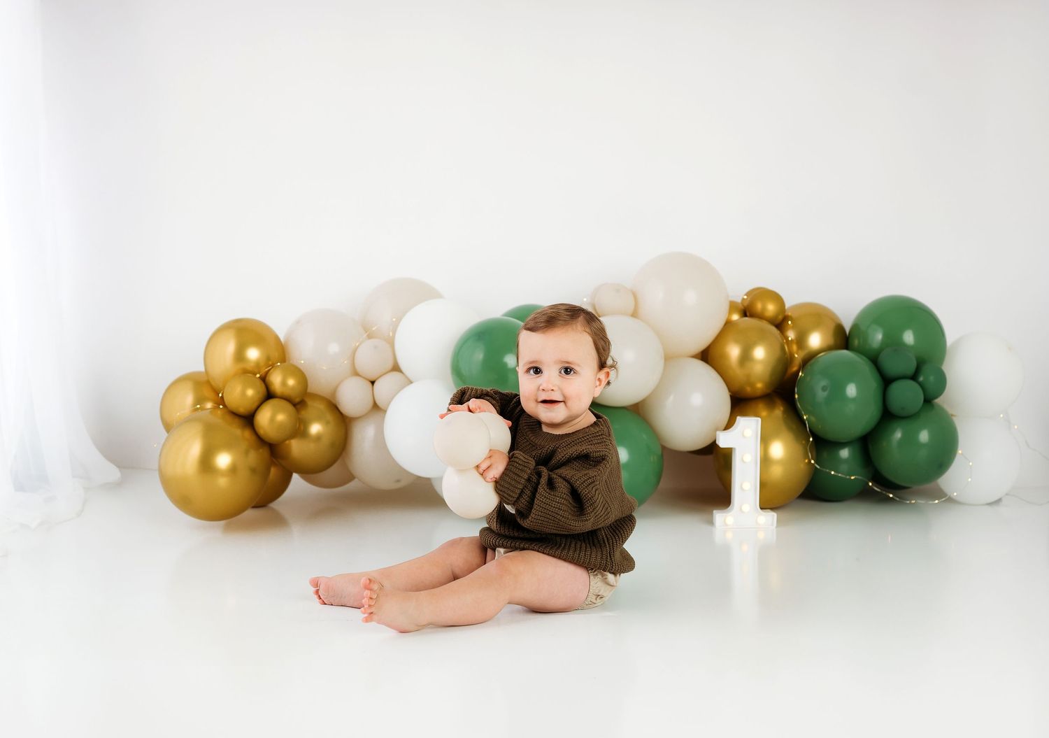 A sleeping baby lies on white fabric surrounded by green, gold, and white balloon decorations.