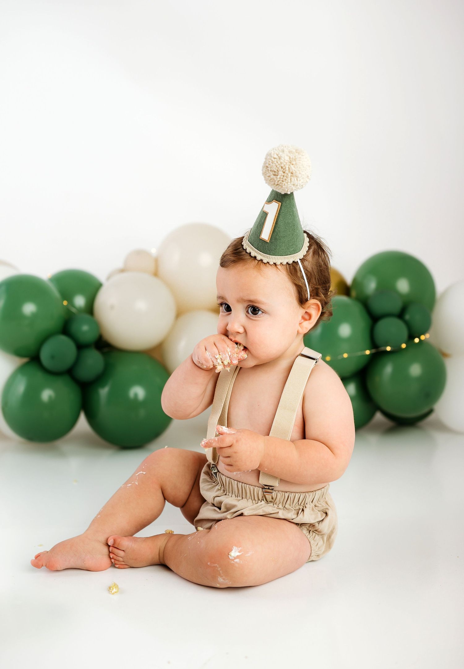 Baby in beige outfit sits among green and white balloons during first birthday cake smash photo session.