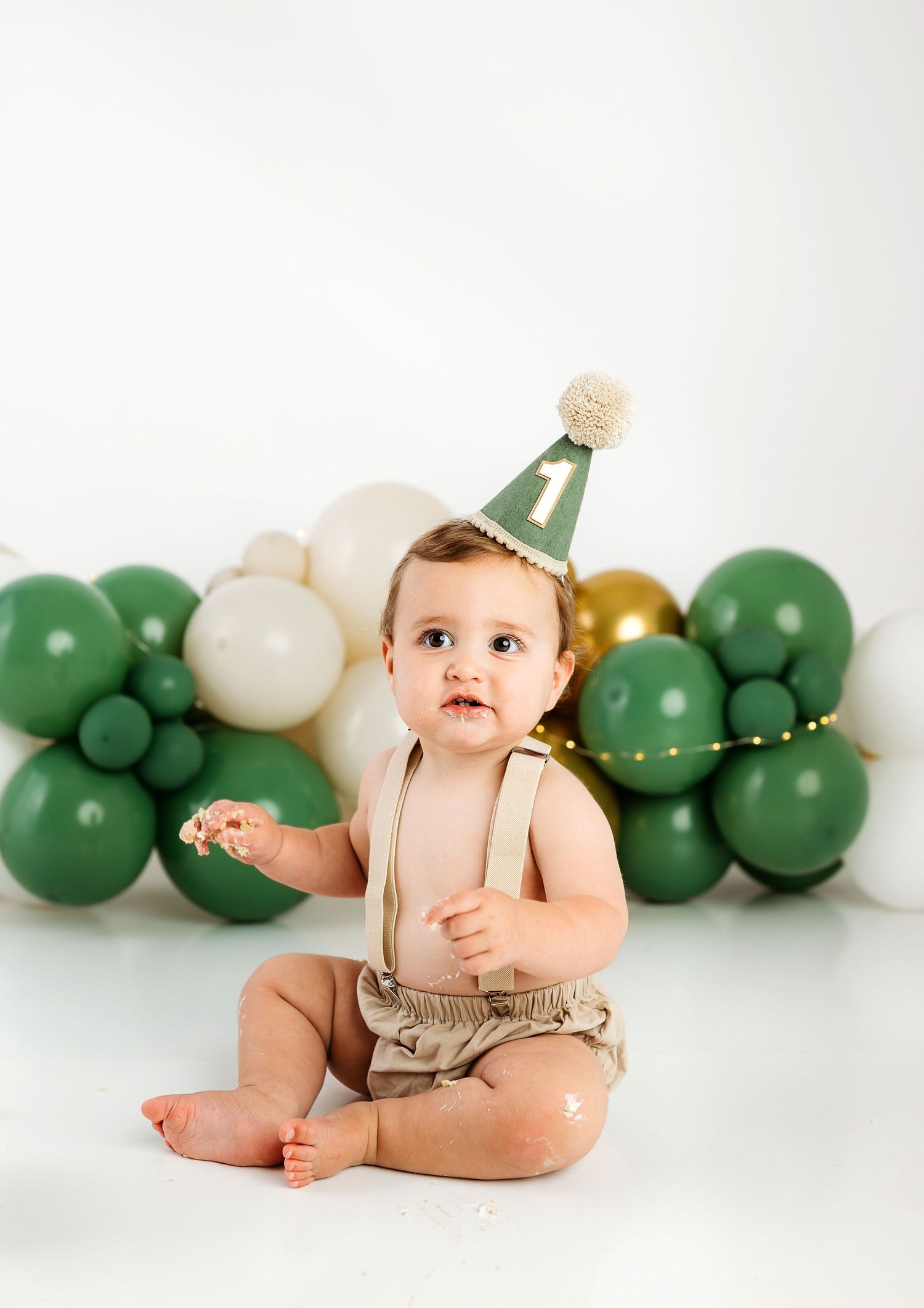 Baby in tan overalls sits on white backdrop with green and white balloon garland decorations.