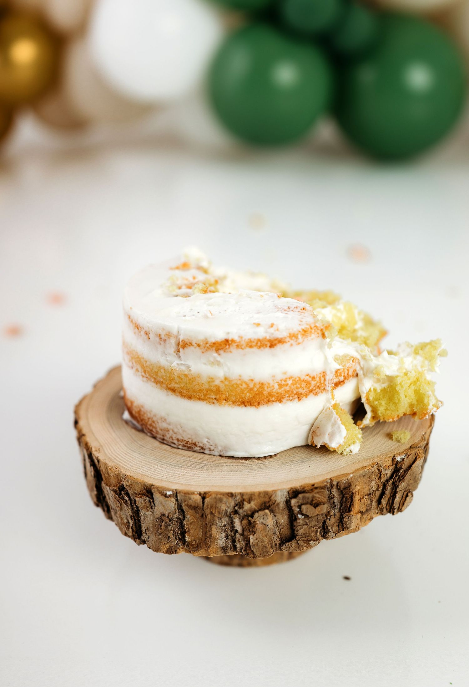 A slice of layered vanilla cake with white frosting sits on a rustic wood slice with green ornaments in the background.