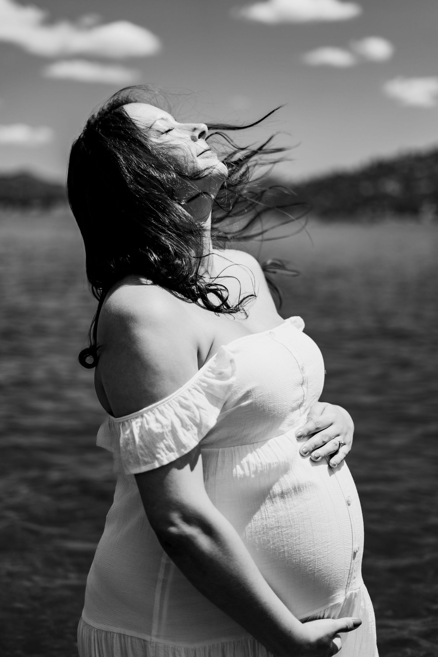 Pregnant woman standing in ocean wearing white dress during black and white maternity session