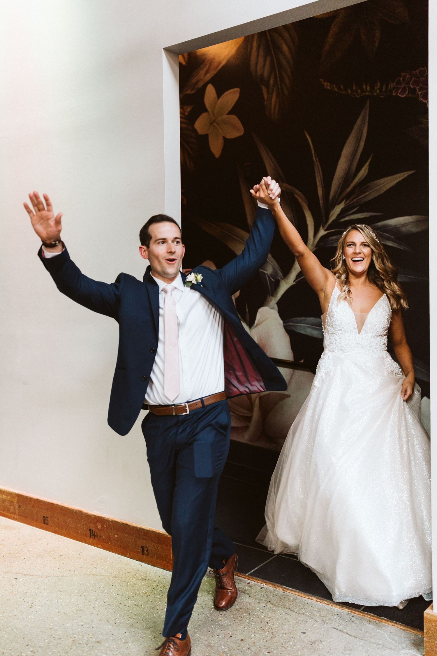 A joyful couple in wedding attire dance and celebrate together in a white-walled venue with dark floral wall decor.