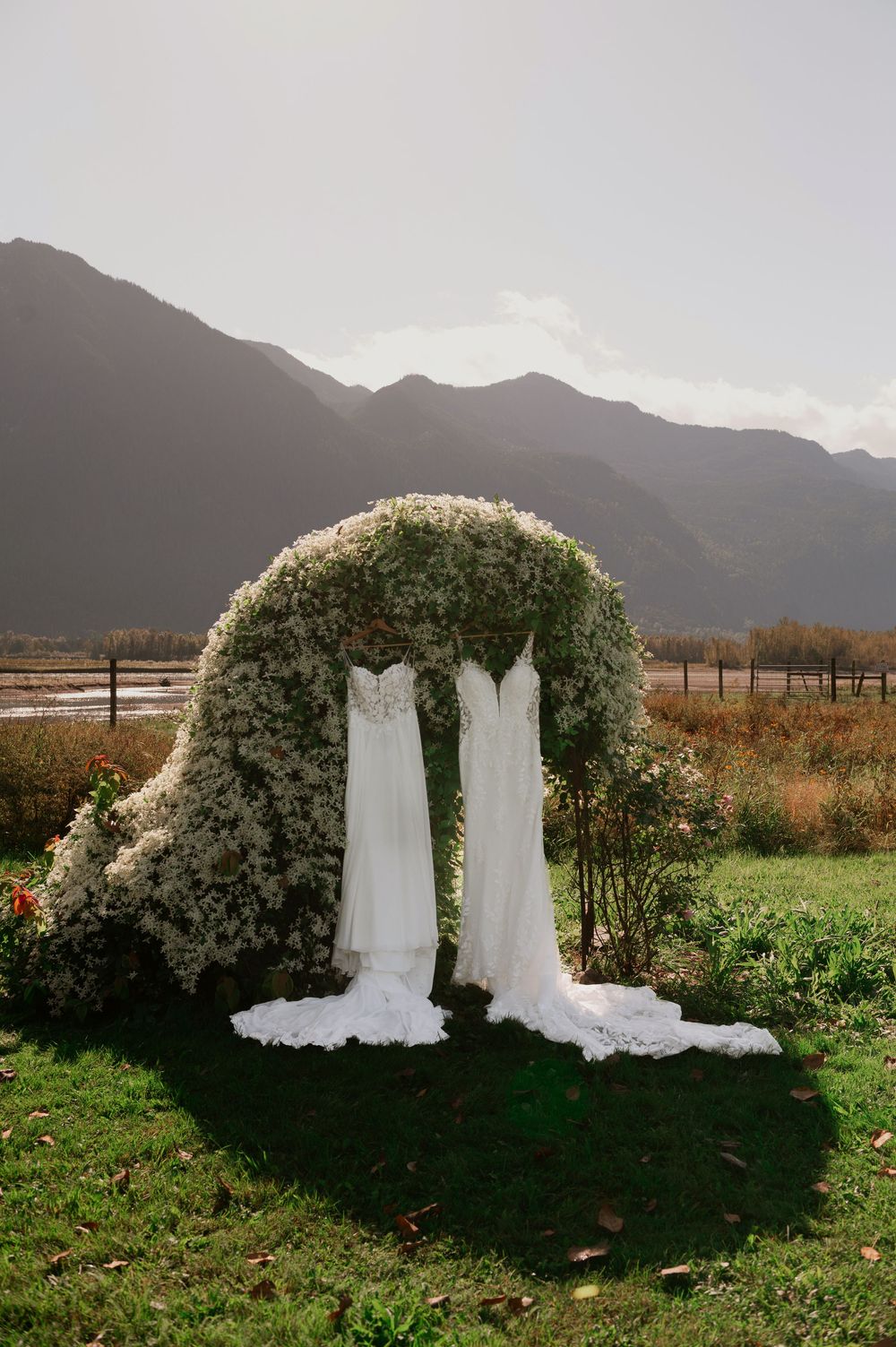 Two white wedding dresses hanging on a floral arch with mountain backdrop.