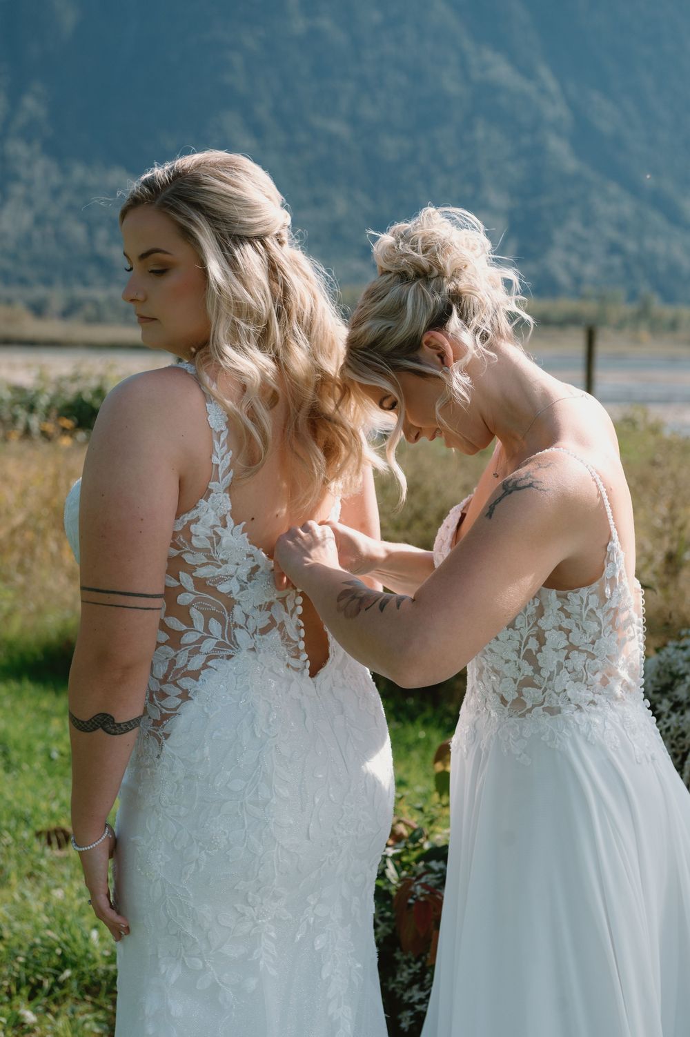 Two people in white lace dresses against mountain backdrop during wedding preparation.