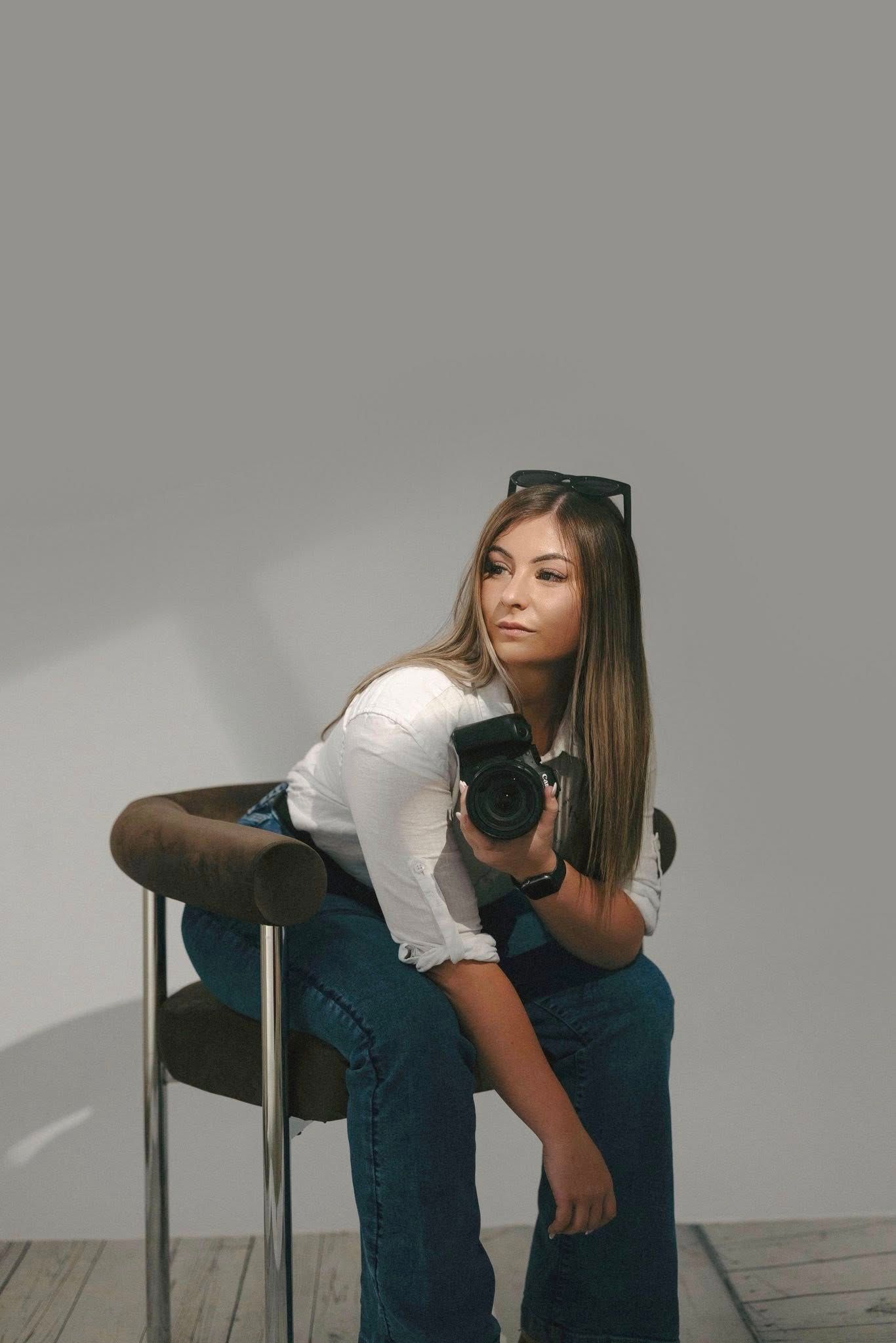 Young female photographer sitting on chair holding DSLR camera with sunglasses on head.