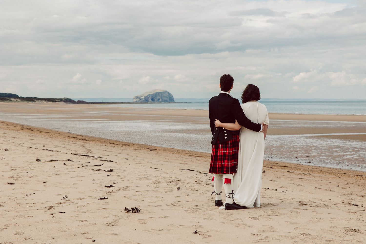 A couple in traditional Scottish attire stands embracing on a scenic beach with mountains visible in the distance.