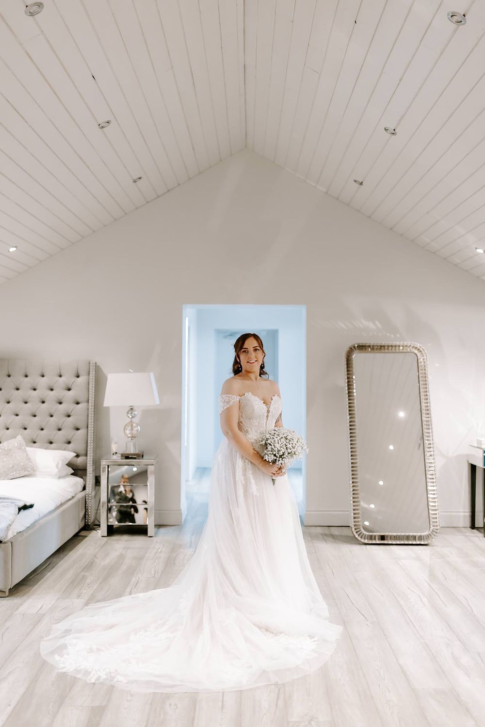 A white wedding dress displayed in an elegant room with vaulted ceilings, gray upholstered headboard, and full-length mirror.