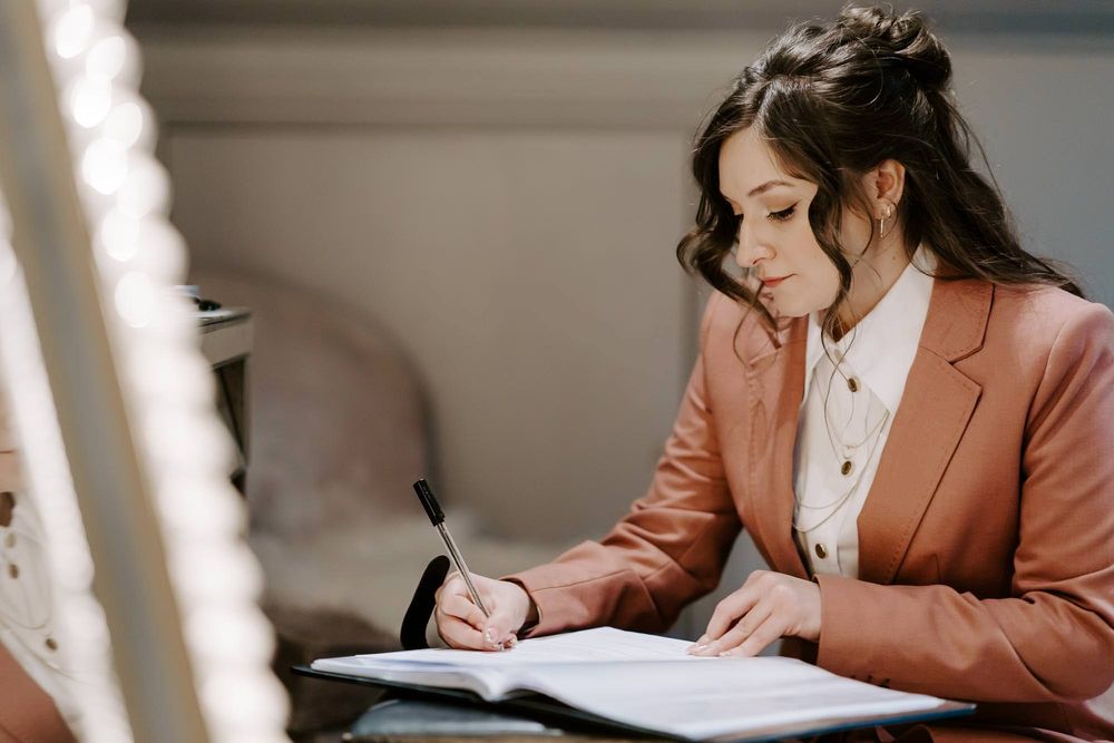 Professional in a peach-colored blazer works at a desk while writing in a notebook.