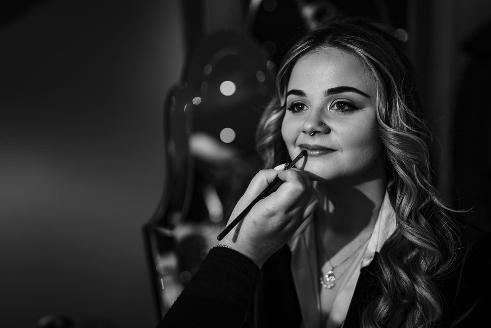 Black and white portrait of someone getting makeup applied with wavy hair and necklace in a dark setting.