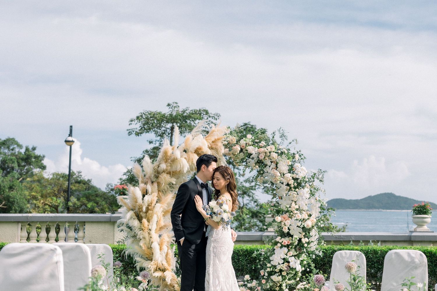 A couple embraces under an elegant floral arch during a waterfront wedding ceremony with ocean views.
