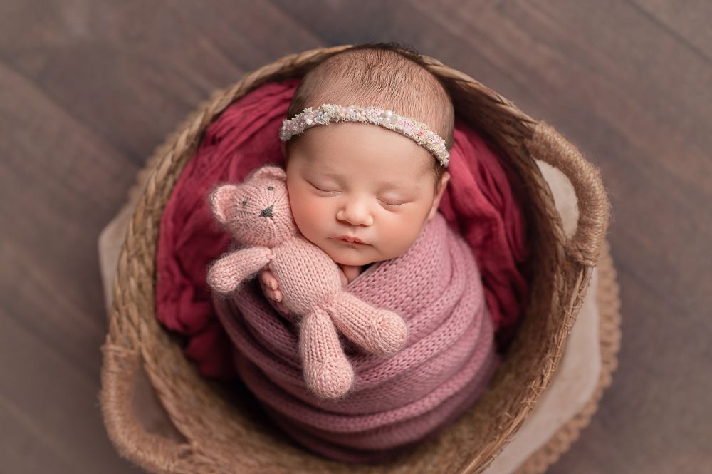 Newborn baby wrapped in pink swaddle sleeping peacefully in round wooden bowl with matching headband accessory.