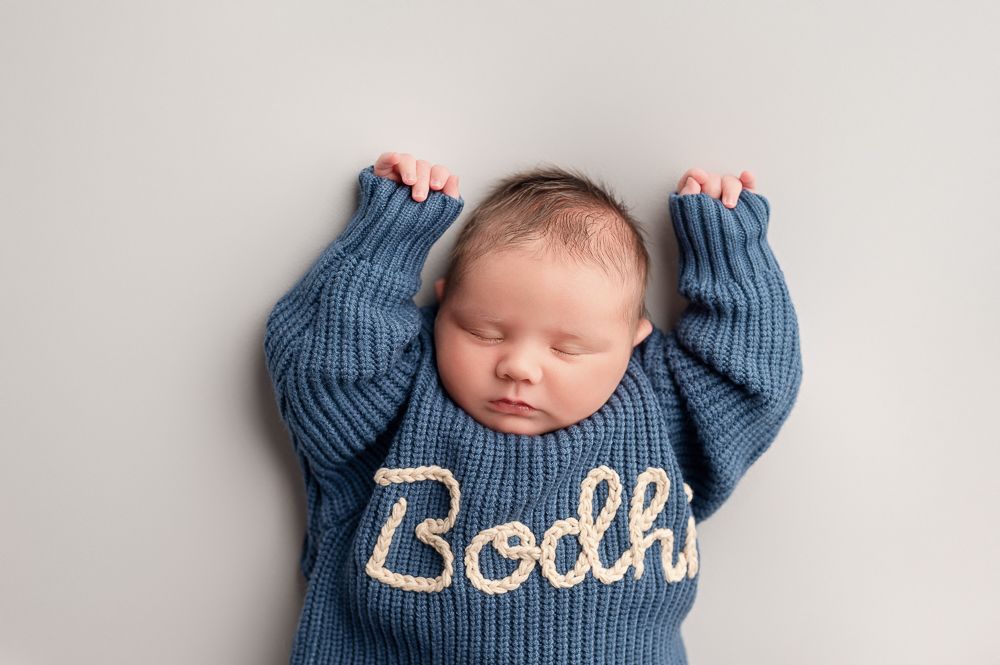Sleeping newborn baby wearing a blue knit sweater lies on white blanket during professional photo session.