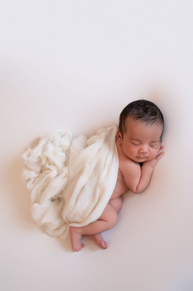 Sleeping newborn wrapped in white fabric lying on light backdrop in minimalist studio setting.