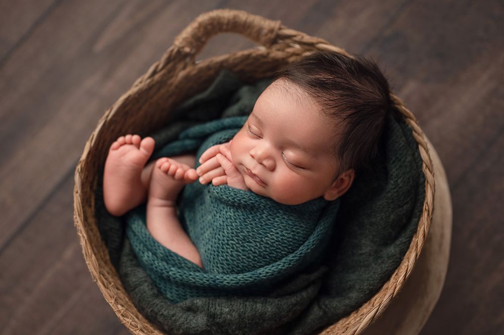Sleeping newborn baby wrapped in teal blanket lying peacefully in rustic wooden basket on dark wood floor.