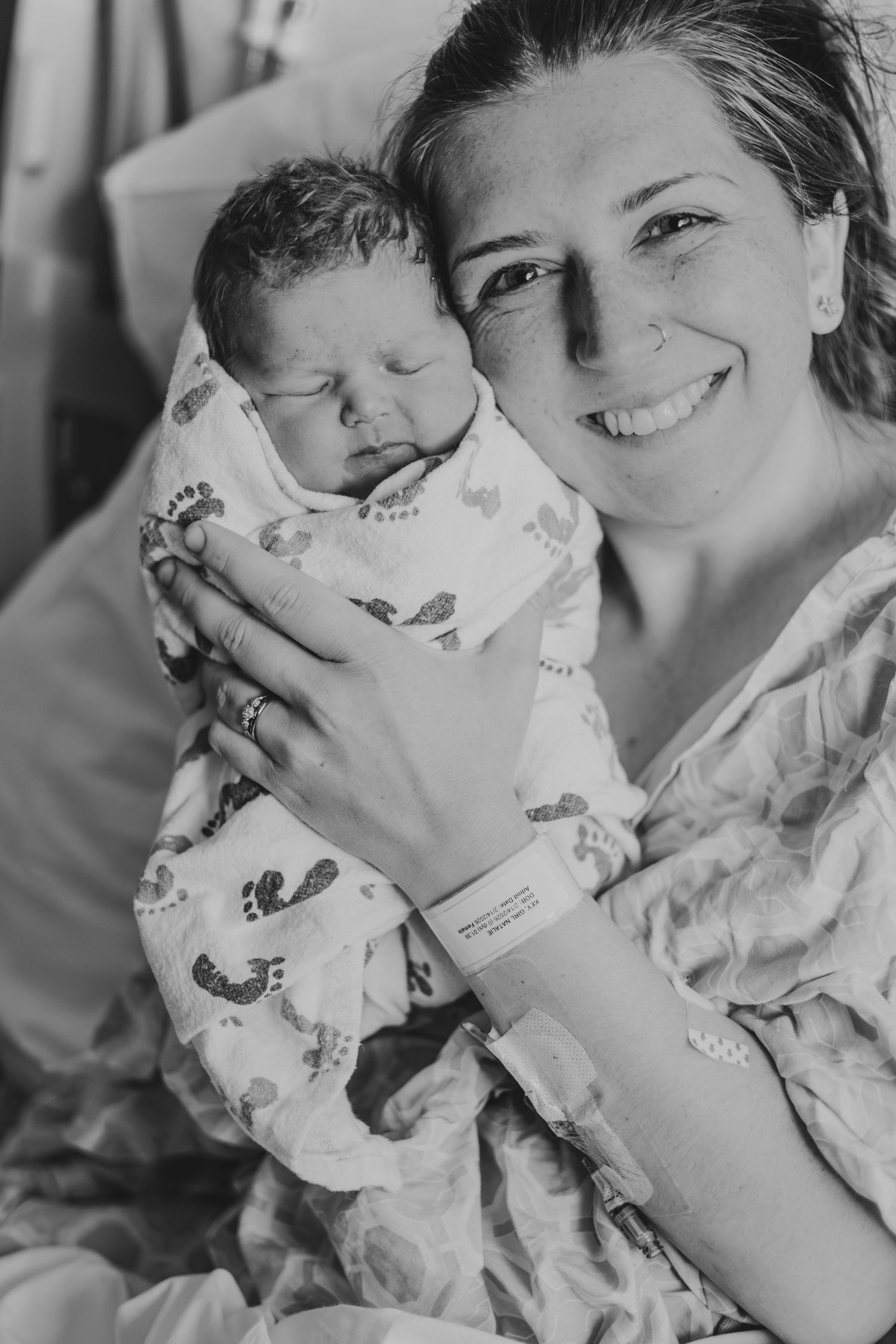 Black and white photo of a smiling new mother in hospital gown holding her swaddled newborn baby | Fresh 48 Hour Photos