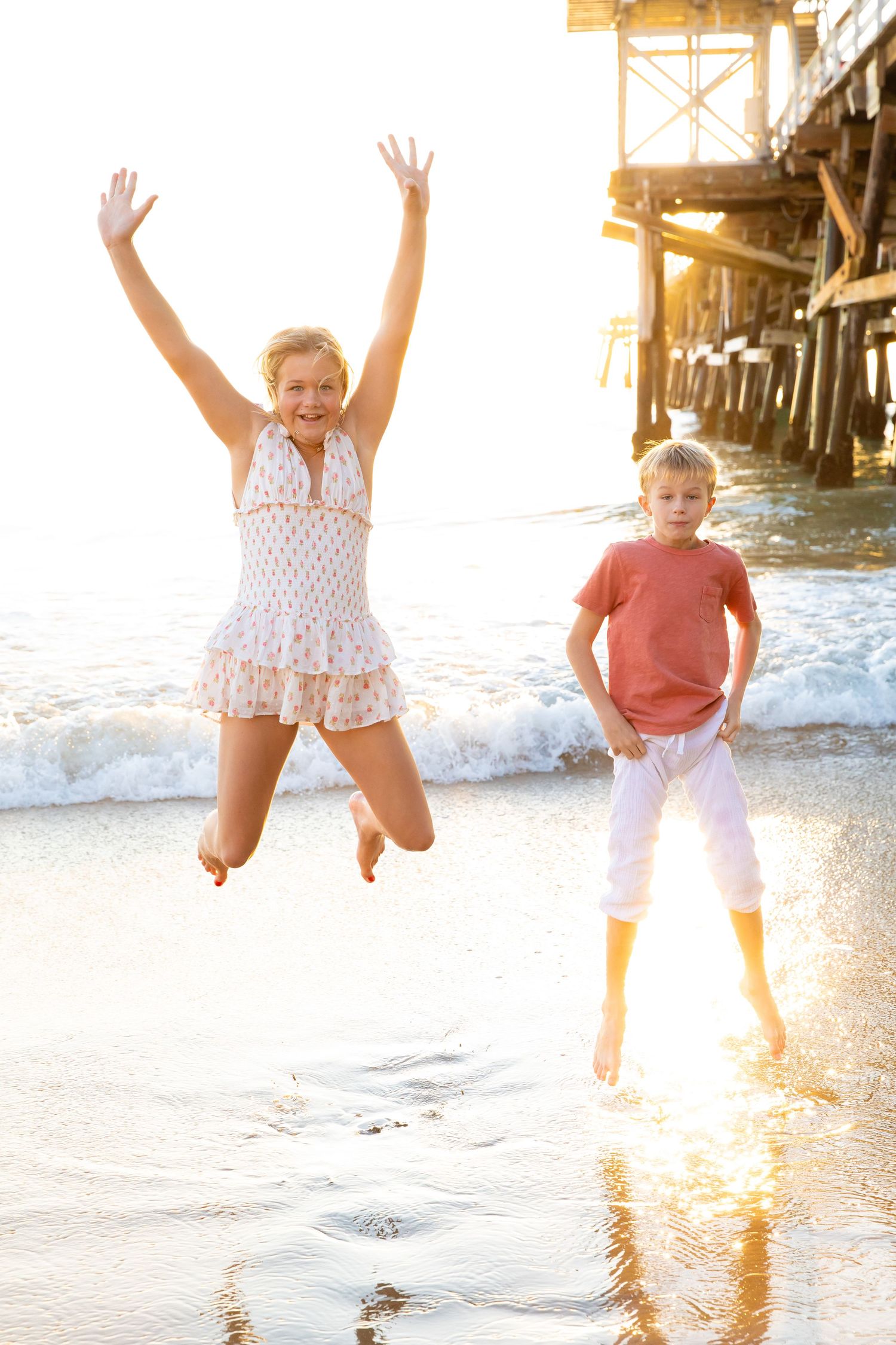 Children jumping and playing at sunset on a beach near a pier with golden sunlight reflecting on the water.