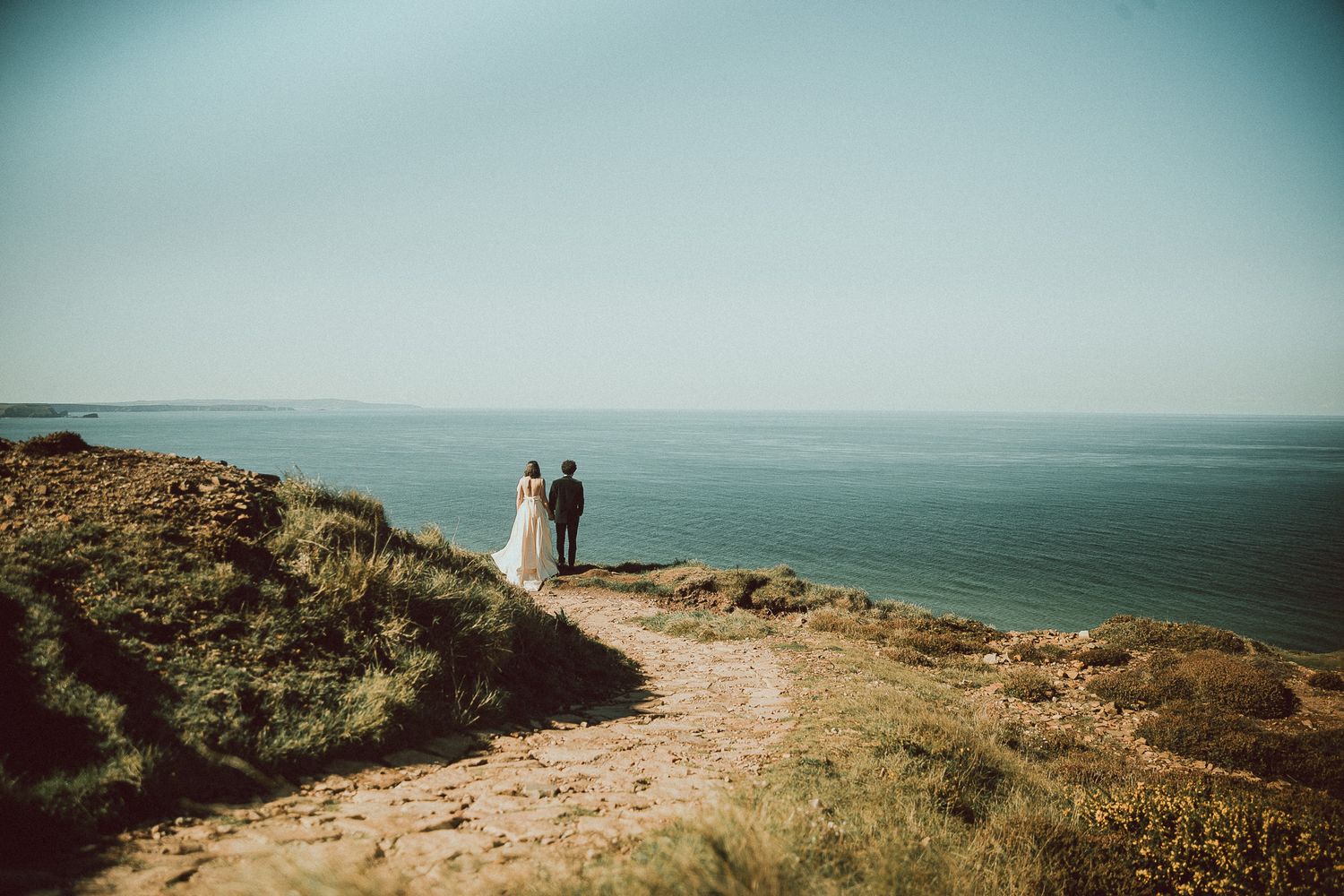 Couple walking along scenic coastal cliffs overlooking the vast blue ocean on a sunny day.