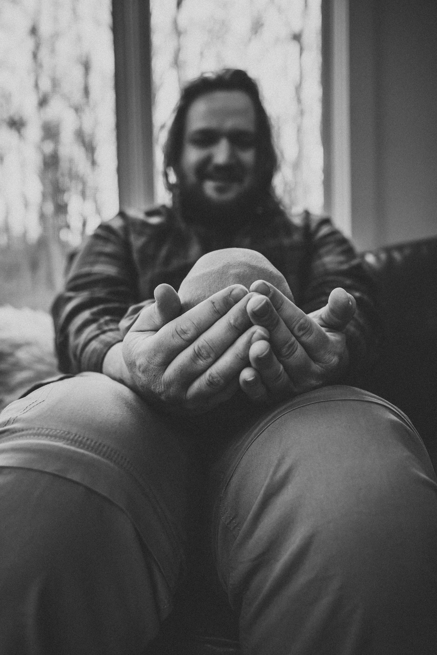 A smiling bearded man cradles a newborn baby in his hands in a black and white portrait photo.
