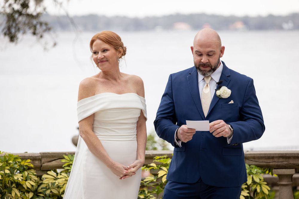 A wedding couple stands on a scenic waterfront terrace with palm trees and water views in the background.