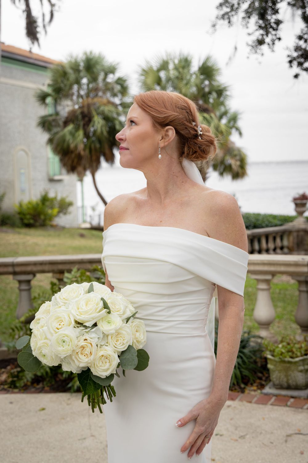 An elegant bridal portrait series featuring an off-shoulder white gown and cream rose bouquet by the water.