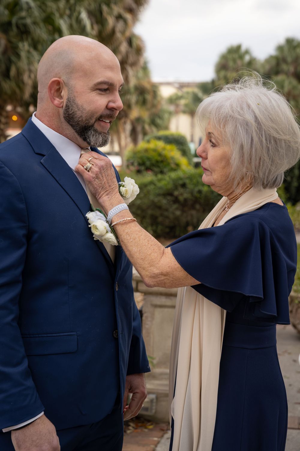 A touching moment between a groom in a navy suit and woman in navy dress adjusting his boutonniere at an outdoor venue.