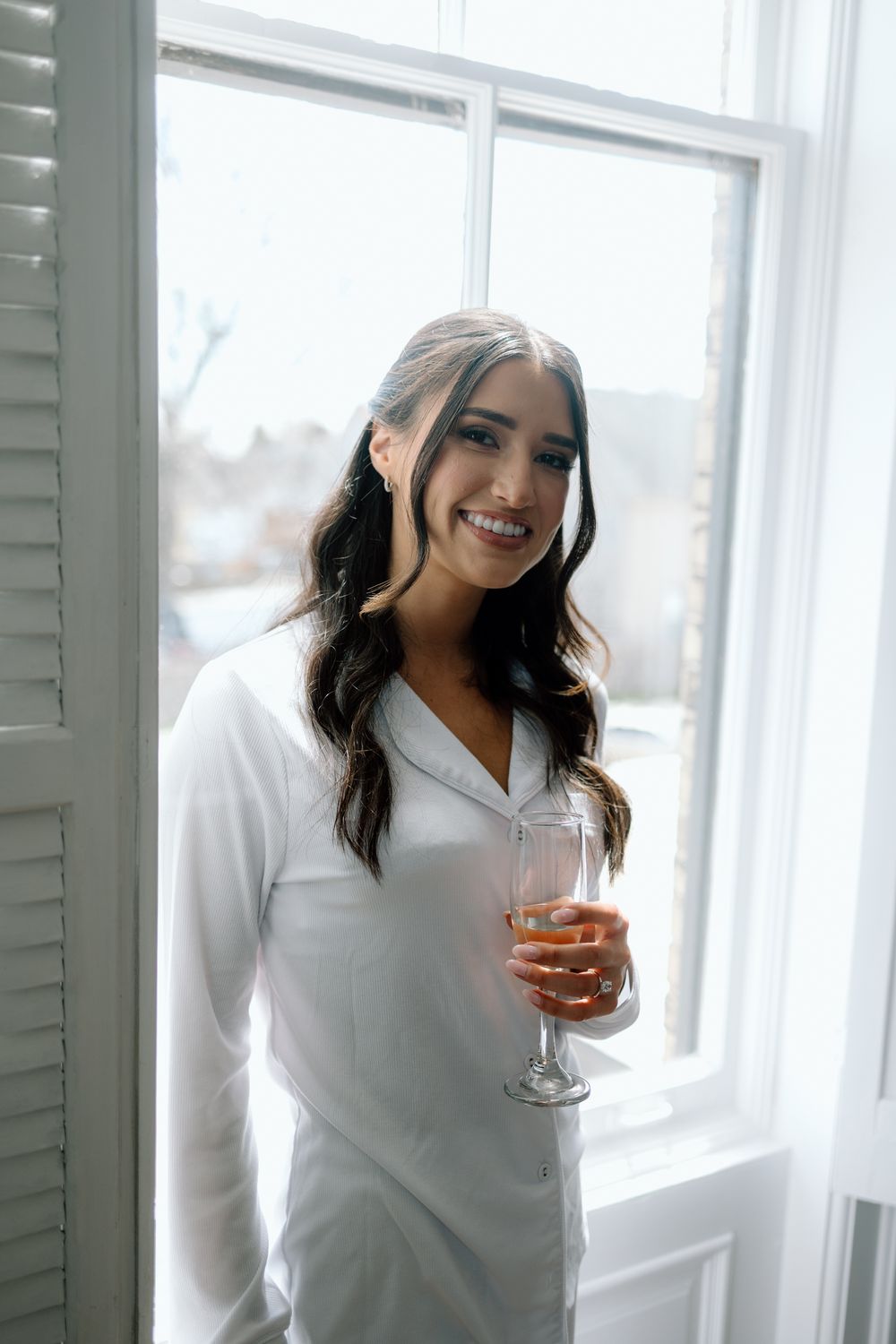 Woman in white sweater smiling while holding wine glass near bright window.