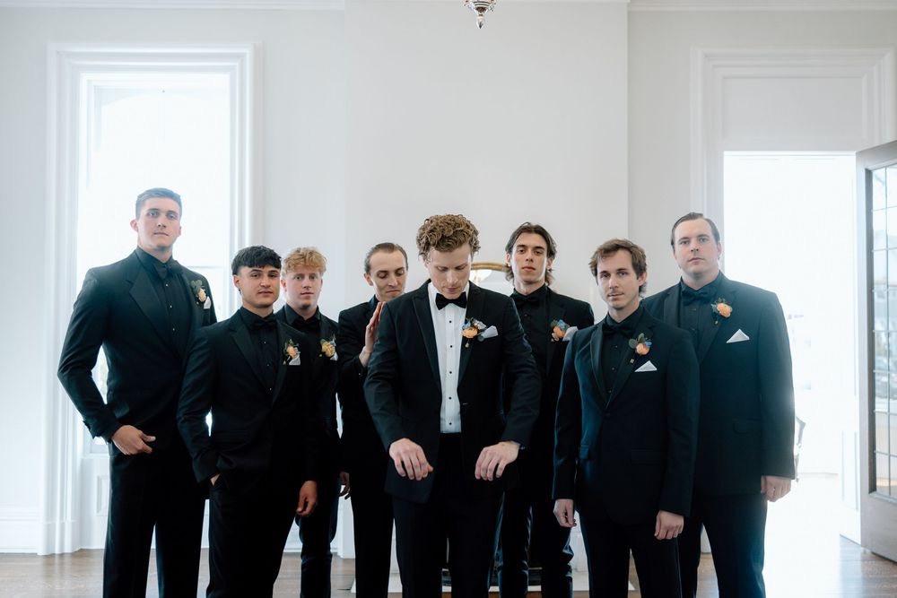 A wedding party wearing black suits and tuxedos poses together in a bright white room.
