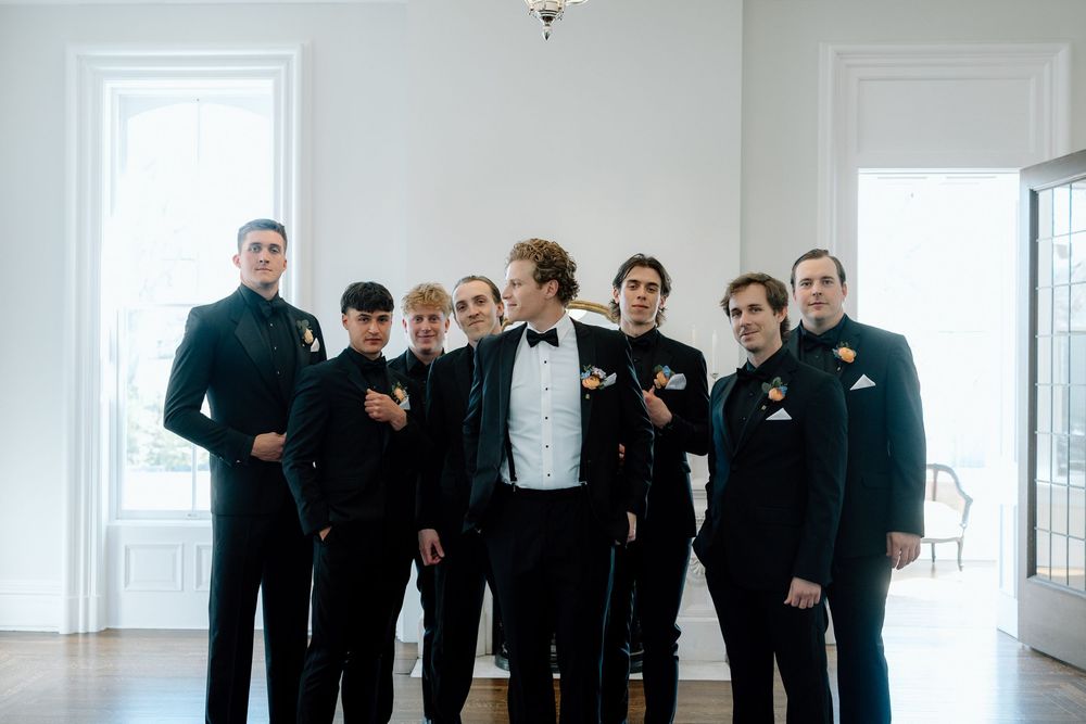 A group of people in formal black attire gather in a bright white room for a wedding party photo.