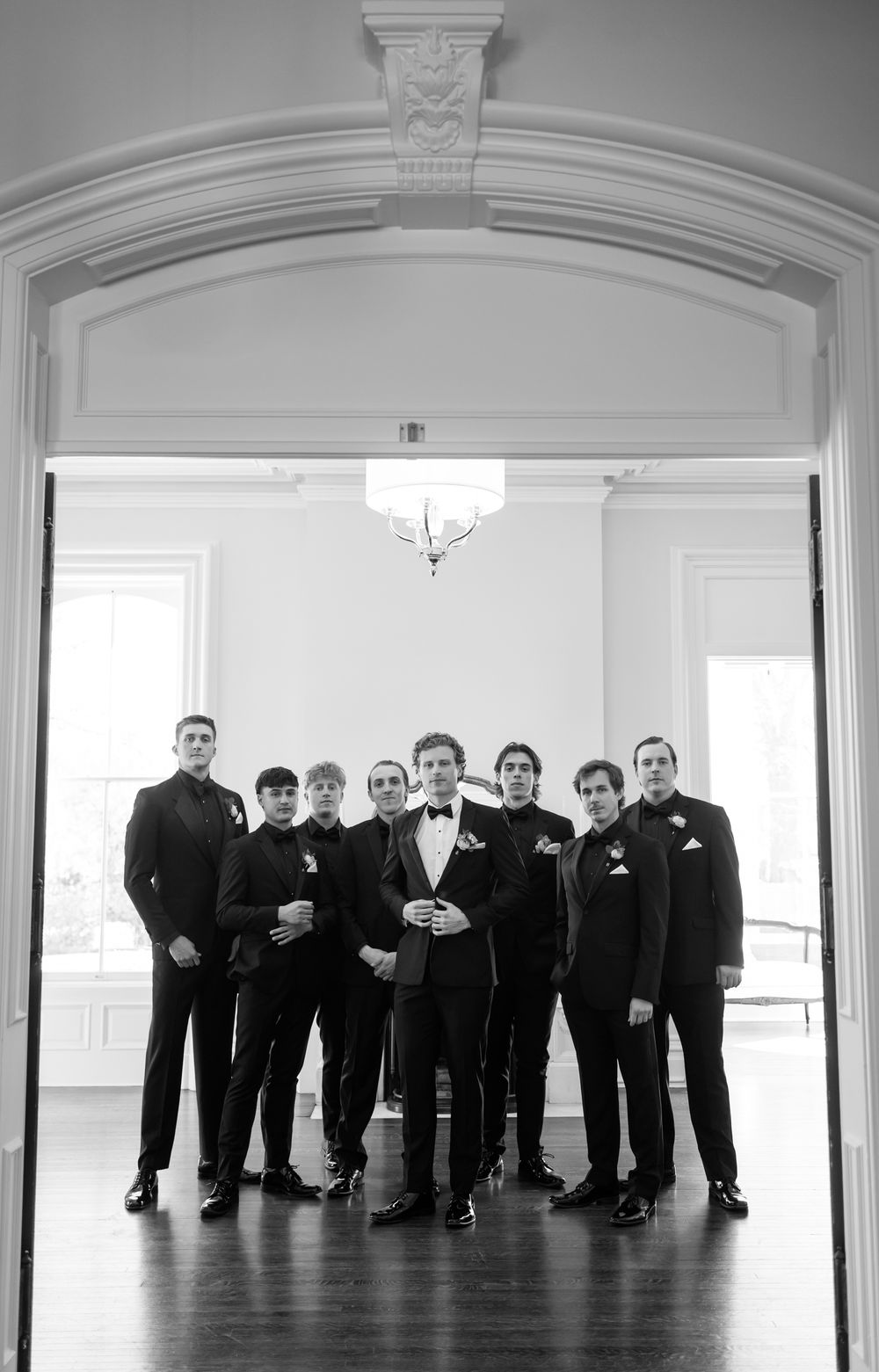 Panoramic black and white shot of groomsmen lined up in elegant room with arched doorways.