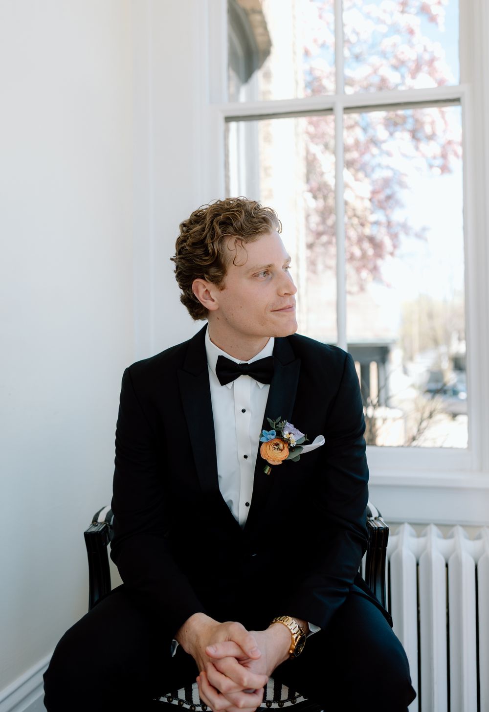 A groom in a classic black tuxedo and bow tie sits thoughtfully by a window with a floral boutonniere.