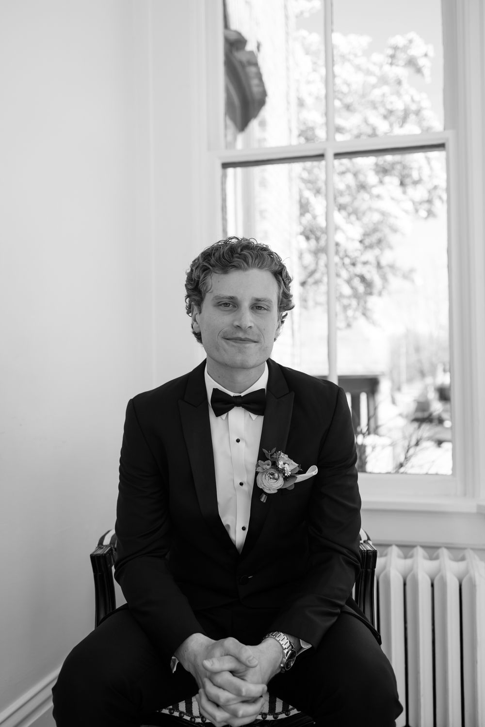 A black and white portrait of a groom seated by a window in formal wedding attire.
