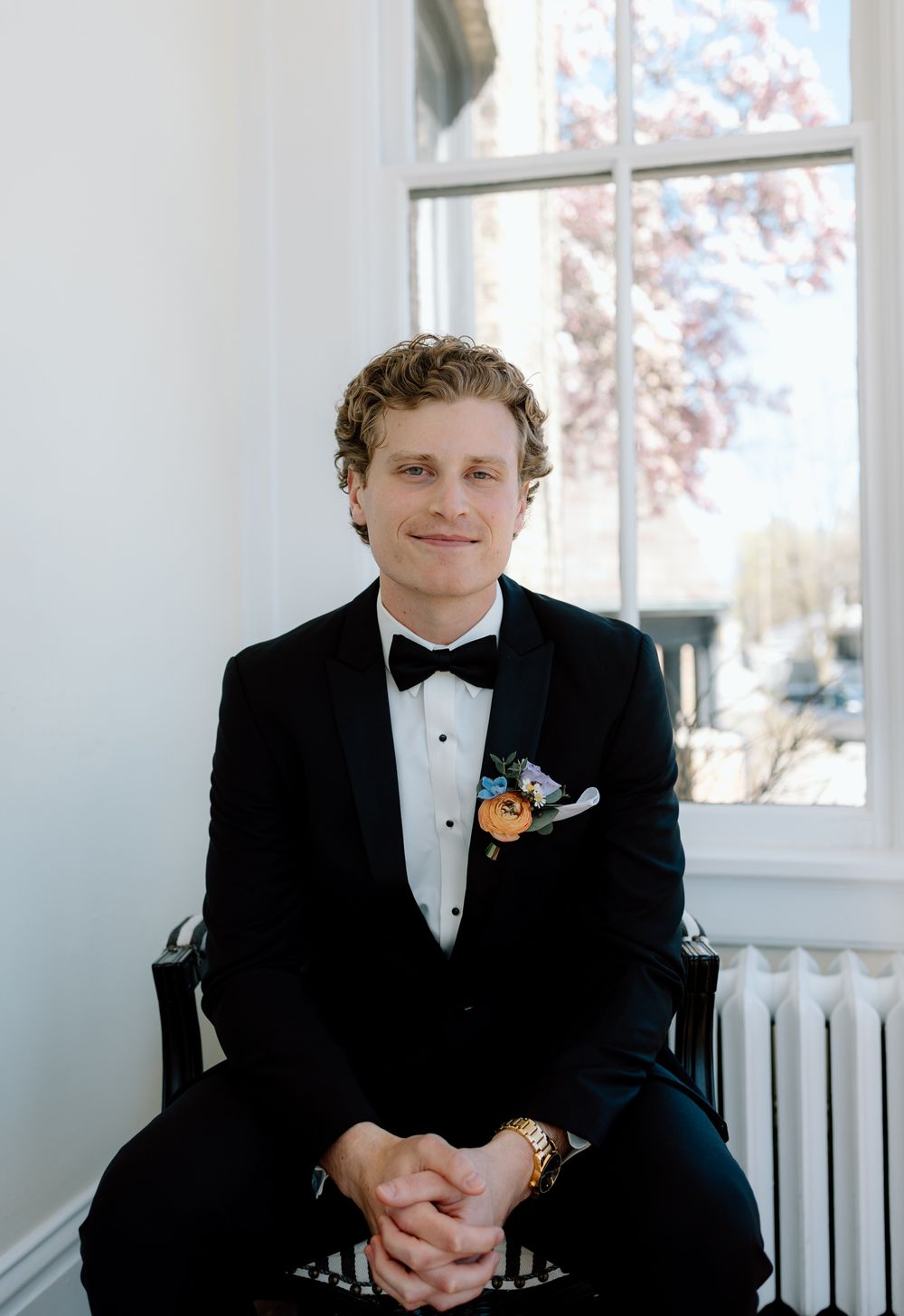 A groom in a black tuxedo and bow tie sits casually in a bright white room by a window.