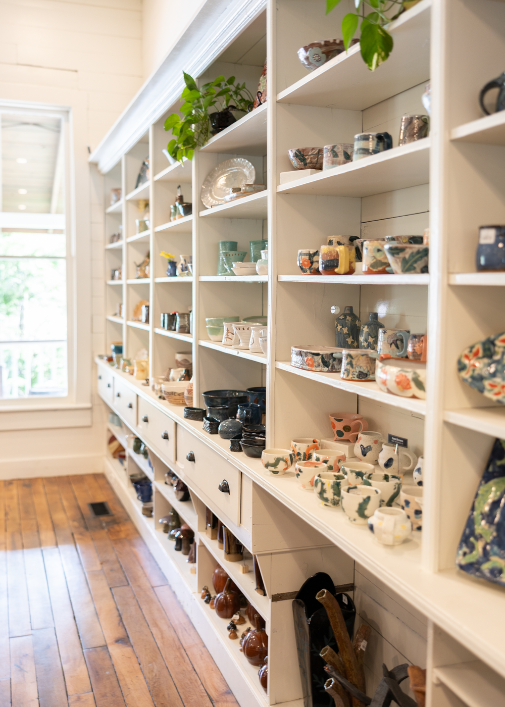 White built-in shelving unit displaying a large collection of handmade ceramic mugs and pottery in a bright shop.