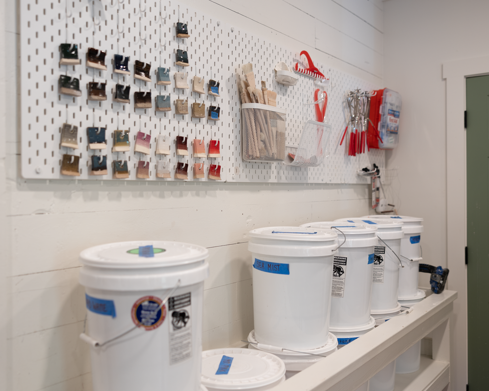 Pottery studio glaze station with white pegboard holding tools and multiple buckets of ceramic glaze.